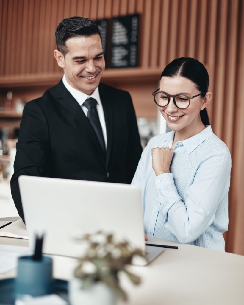 Two smiling young businesspeople working on a laptop and going over notes while standing together at a table in an office